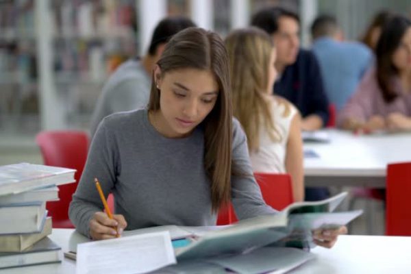 Focused female young student at the library reading books and taking notes with a pencil on her notebook